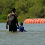 (१ अगस्ट २०२३ मा टेक्सासको इगल पासस्थित रियो ग्रान्डे नदीमा सीमा अवरोधका रूपमा प्रयोग गरिएका ठूला 'बुई' (पानीमा तैरिने साधन) हरूको छेउबाट आप्रवासीहरू हिँड्दै गरेको दृश्य। एपी फाइल तस्बिर: एरिक गे)